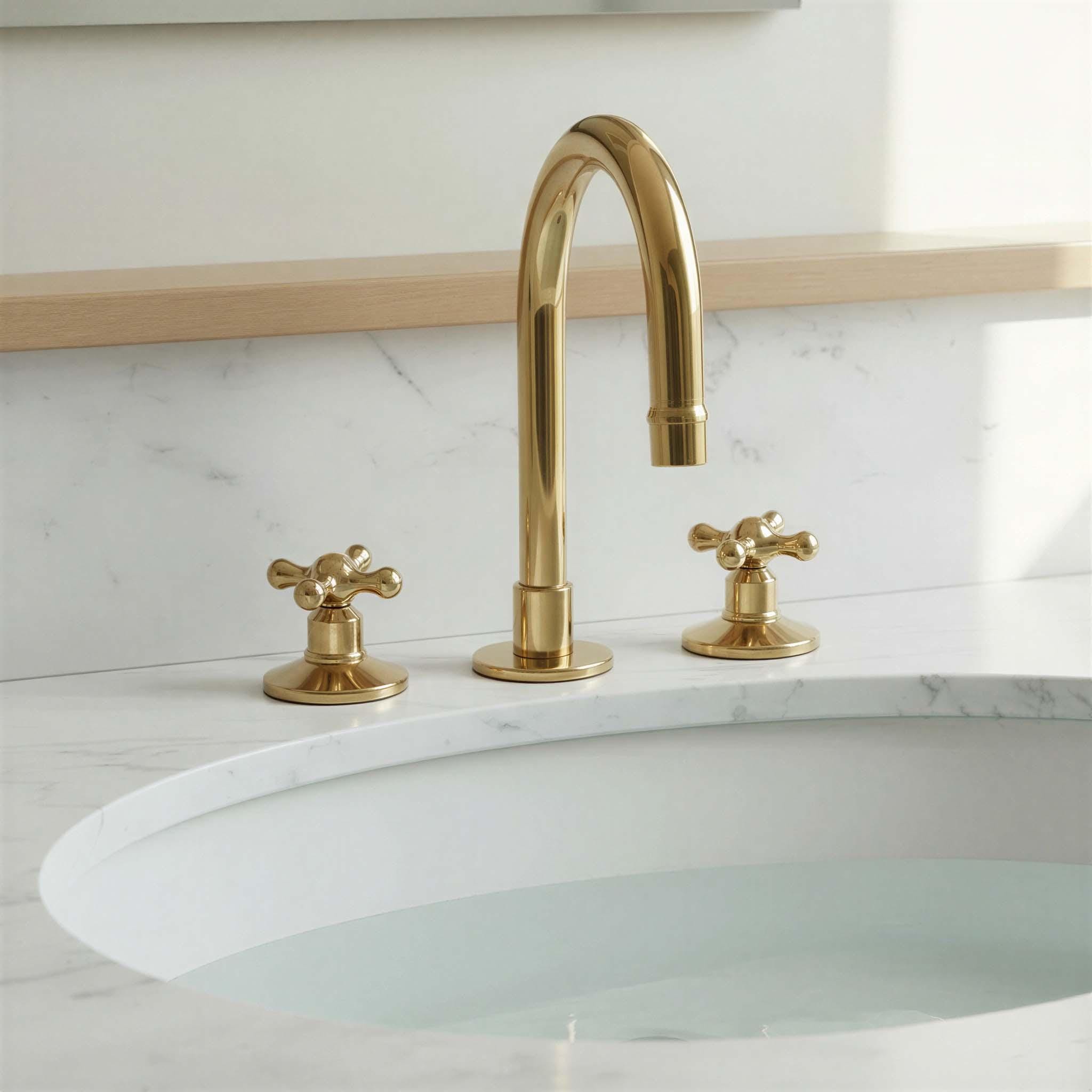 Gold faucet with two handles on a white sink against a light marble wall.