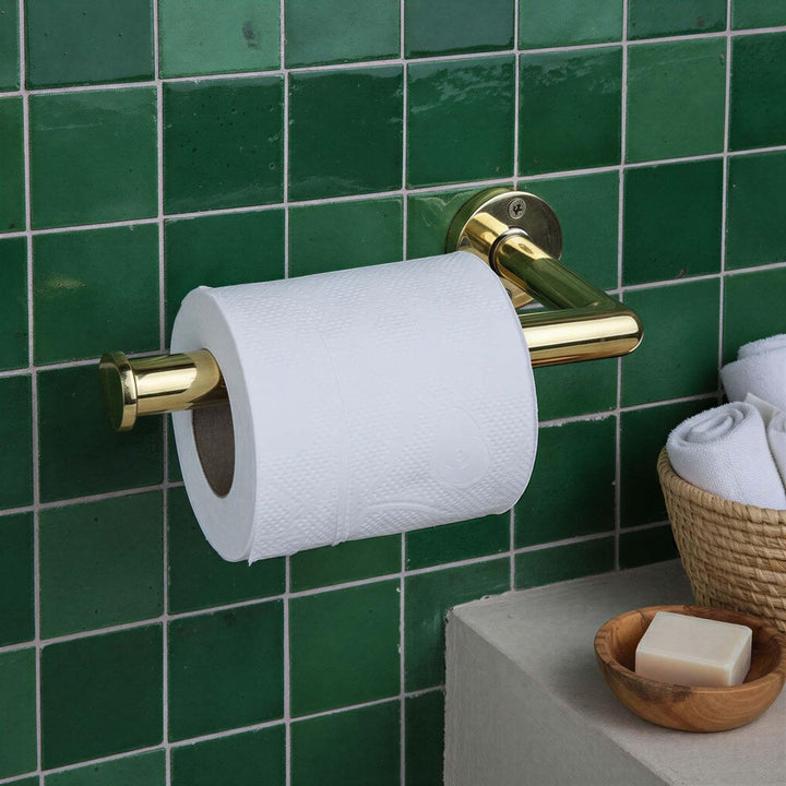 Gold toilet paper holder with a roll of toilet paper against a green tiled wall.