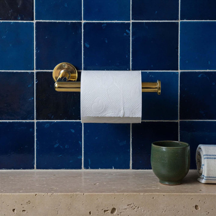 Gold toilet paper holder with a roll of toilet paper against a blue tiled wall.