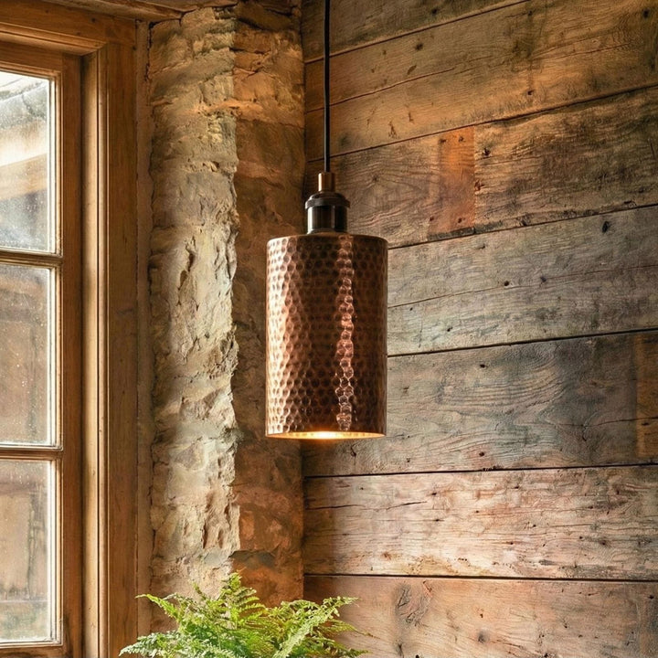 Copper pendant light hanging against a rustic wooden wall with a window in the background.
