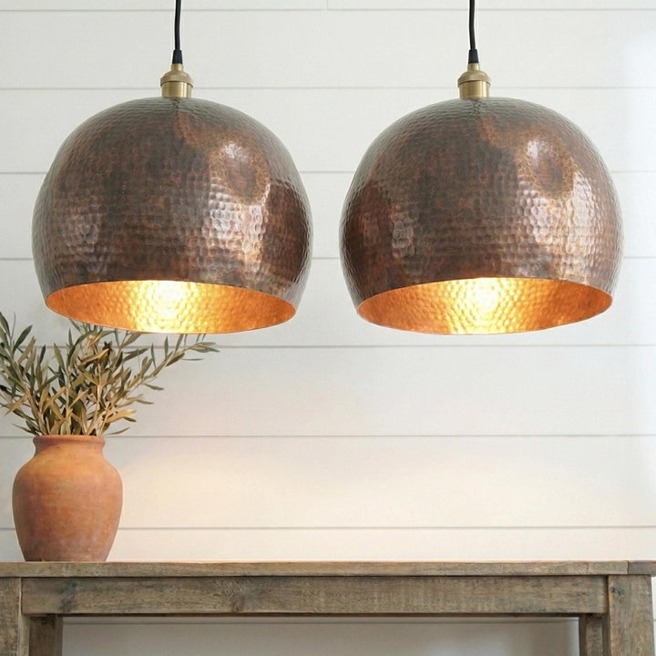 Two copper pendant lights hanging above a wooden table with a plant on a white paneled wall background.