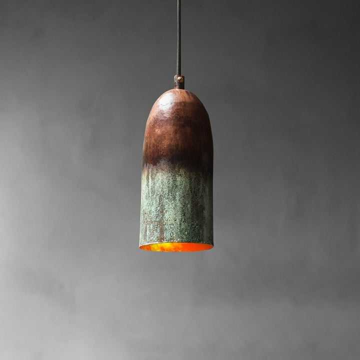 A single pendant light with a bullet-shaped shade made of oxidized metal, showing a dark brown top fading to a greenish patina at the bottom, with a glowing orange interior, suspended against a dark gray background.