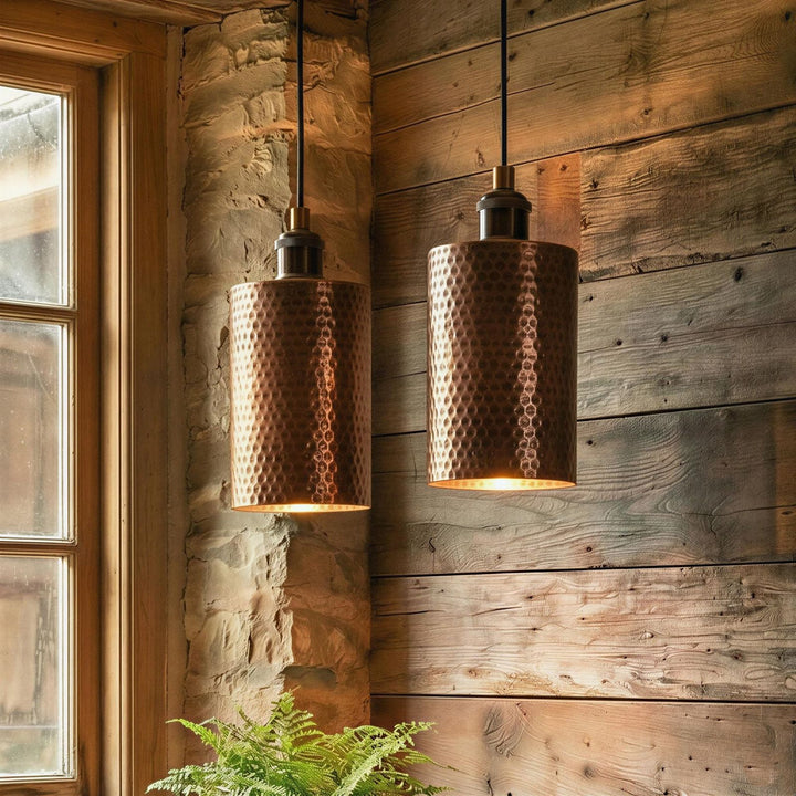 Two copper pendant lights hanging against a wooden wall with a window and plant in the foreground.