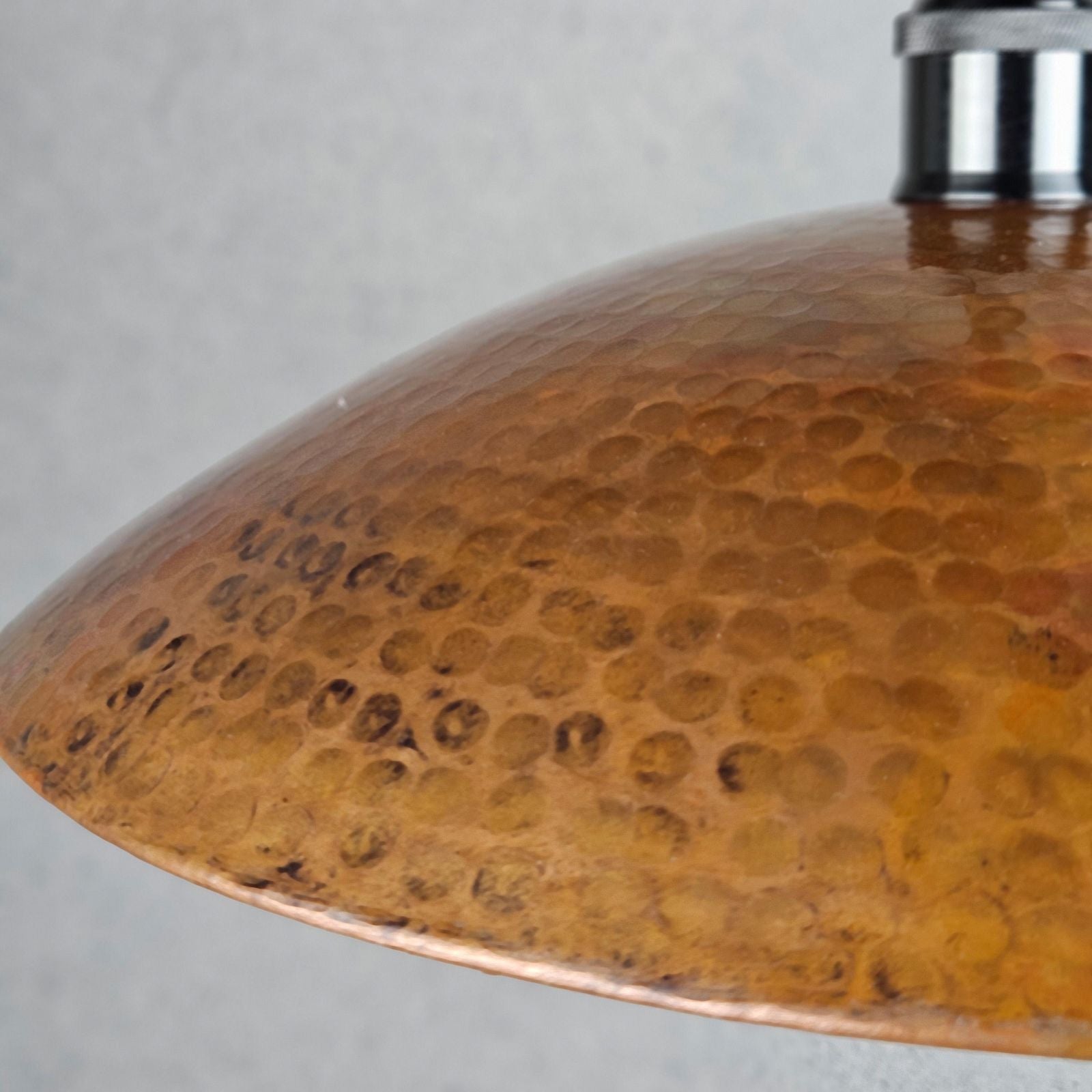 Close-up of a hammered metal dome shade in warm coppery gold tones, with a small portion of a dark metal fixture visible at the top against a light gray background.