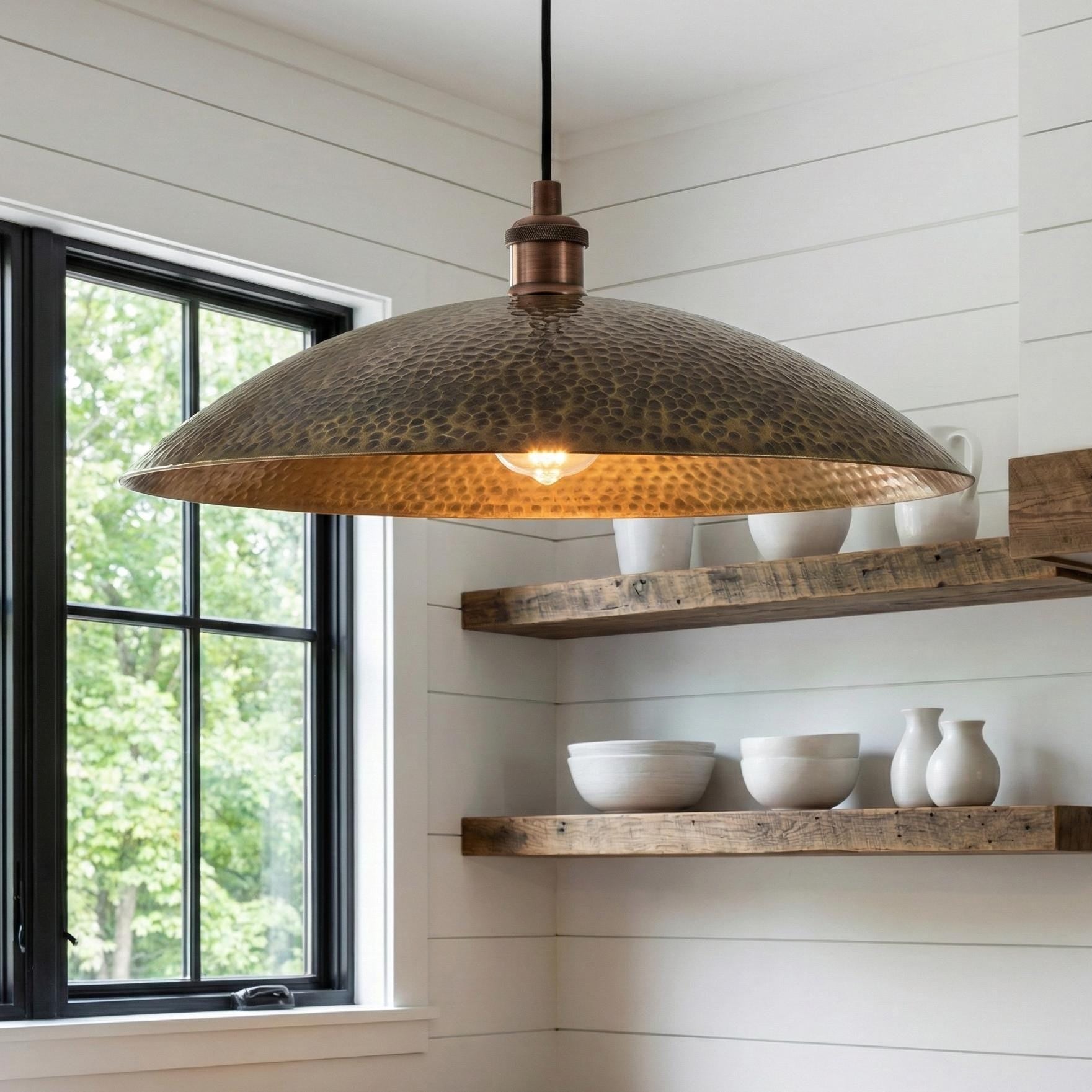 Hammered metal pendant light hanging in a kitchen with wooden shelves and windows.