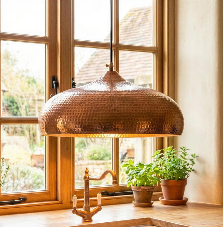 Copper pendant light hanging in a room with large windows and potted plants on a windowsill.