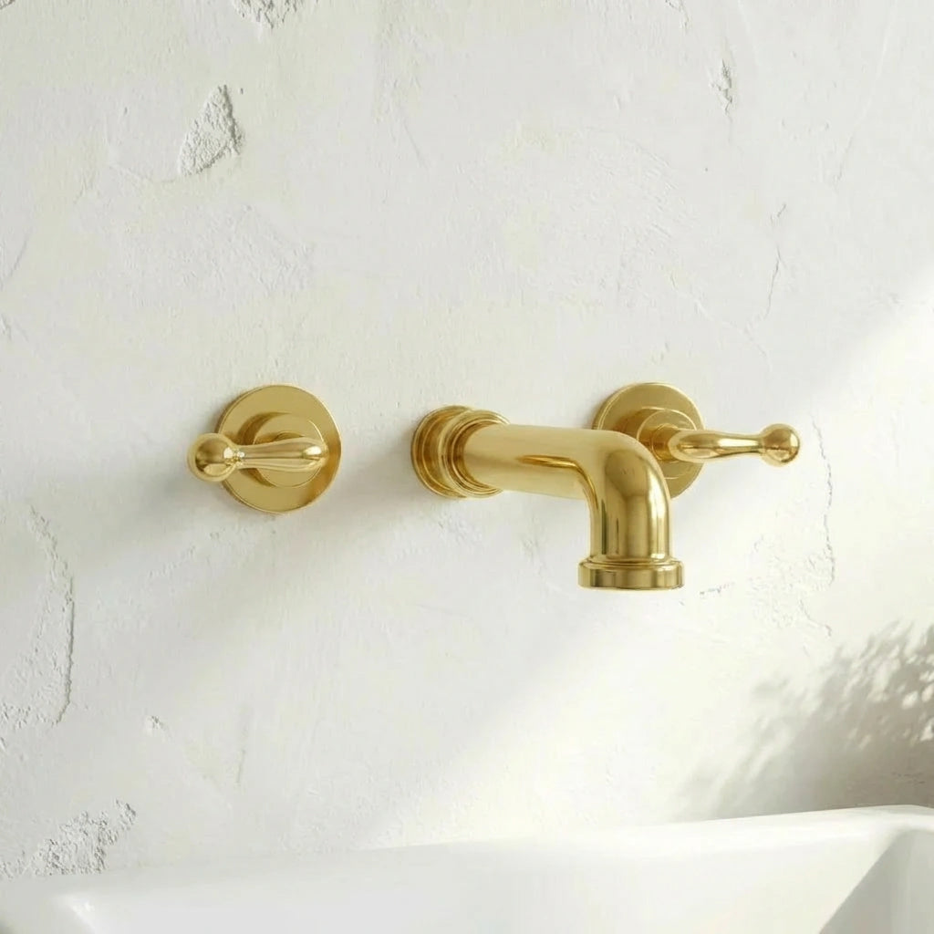 A polished brass wall-mounted bathroom faucet with two matching lever handles, installed on a textured white plaster wall above a white sink.