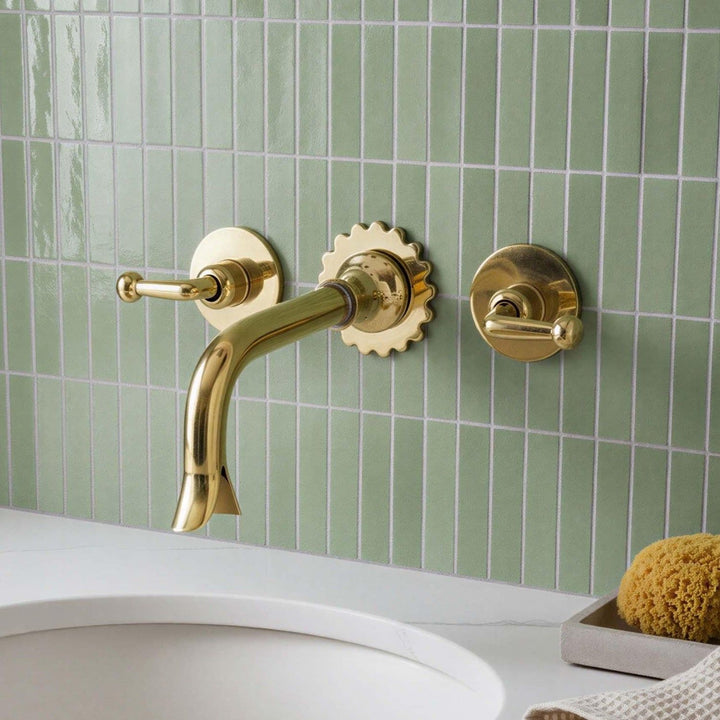 Gold faucet and handles on a green tiled wall with a sink and sponge in the foreground.