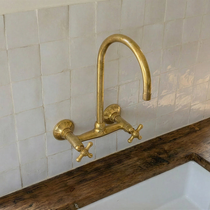 A polished brass wall-mounted kitchen faucet with a high-arc spout and two cross handles, installed above a white sink on a rustic wooden countertop against a white tiled backsplash.