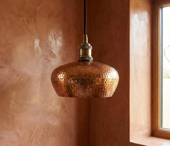 Copper pendant light hanging against a textured brown wall with natural light coming through a window.
