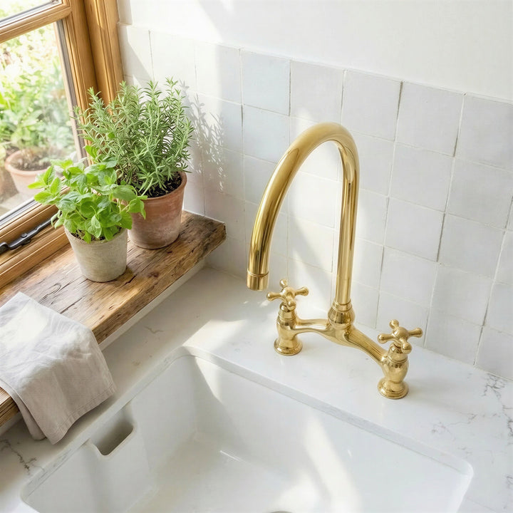A polished brass bridge faucet with two cross handles installed in a white farmhouse sink, set against a white subway tile backsplash with a wooden window sill holding potted green plants.
