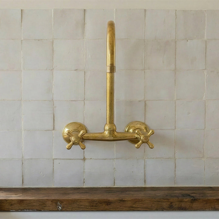 A vintage-style brass wall-mounted faucet with two cross handles, installed above a dark wood sink against a white tiled backsplash.